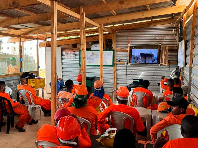 Trabalhadores uniformizados de laranja, com capacetes, assistem a um treinamento em vídeo em um canteiro de obras.